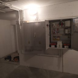 Eletrik - Otwarta szafa sterownicza z widocznymi elementami instalacji elektrycznej, przewody w białych rurkach na ścianie, akumulatory na dolnej półce.