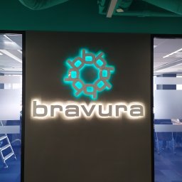 Eletrik - Podświetlane logo 'bravura' na szarej ścianie w biurze. Logo w kolorze turkusowym, nazwa firmy biała. Widoczne rury wentylacyjne pod sufitem i fragmenty biura za szybą.