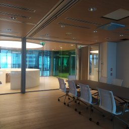 Eletrik - Nowoczesna sala konferencyjna z drewnianą podłogą i sufitem, szklanymi ścianami, oświetleniem sufitowym i stołem z krzesłami. Widoczne znaki ewakuacyjne.