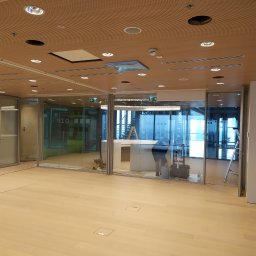 Eletrik - Montaż oświetlenia sufitowego w biurze w Gzy. Widoczne otwory na lampy i pracownik podczas instalacji w przeszklonym pomieszczeniu. Jasna podłoga i drewniany sufit.