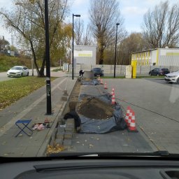 Eletrik - Układanie kostki brukowej przy chodniku, widoczne narzędzia i stożki ostrzegawcze. Prace ziemne w toku, teren zabezpieczony. Jesienne otoczenie, widok z wnętrza pojazdu.