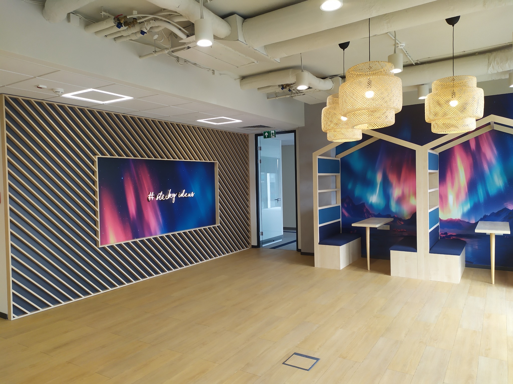 Nowoczesne biuro z drewnianą podłogą, ścianą z ekranem wyświetlającym napis '#sticky ideas' i ażurowymi lampami nad strefą relaksu z boksami.