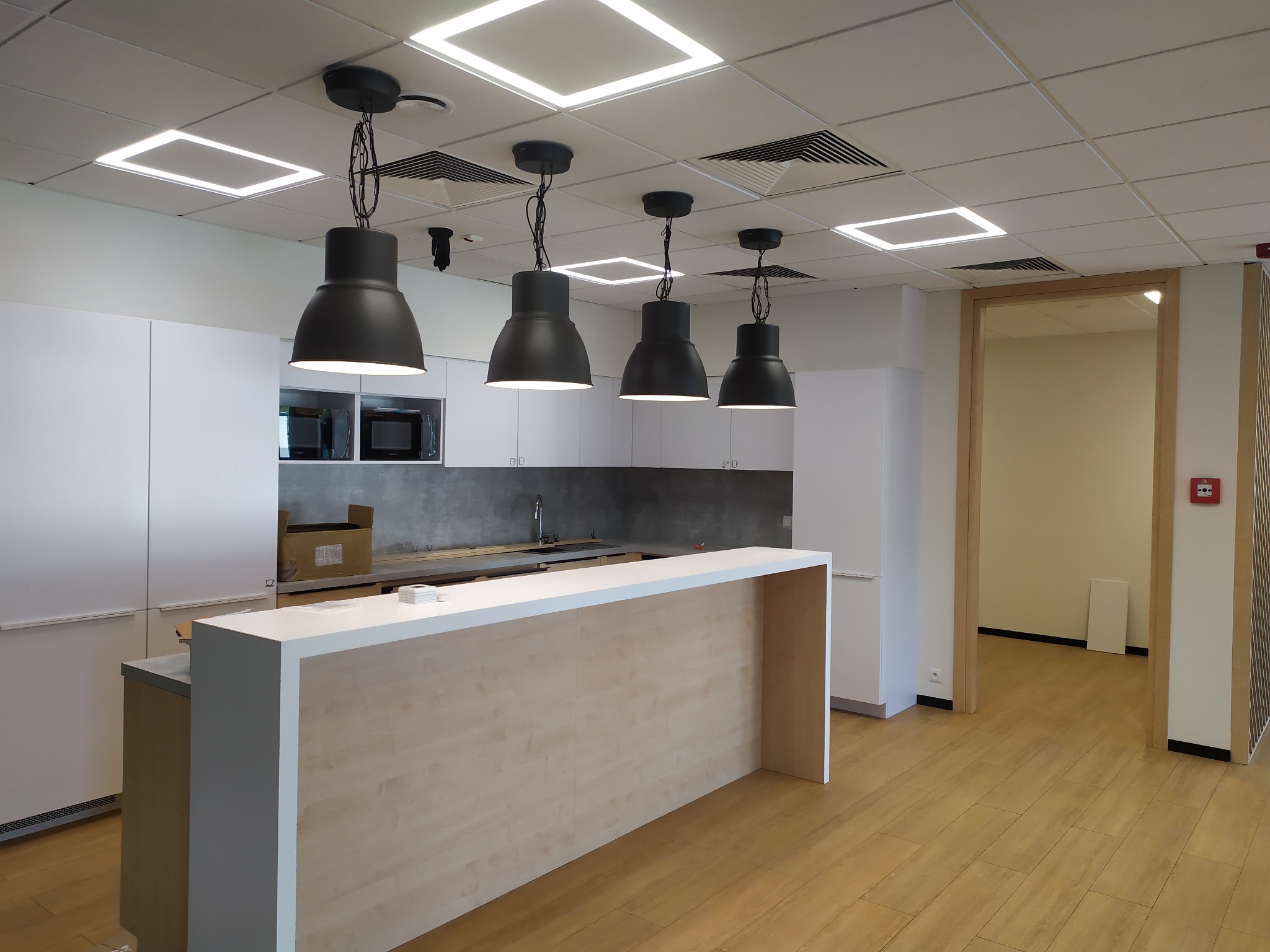 Nowoczesna kuchnia biurowa z białymi szafkami i wyspą. Nad nią cztery czarne, industrialne lampy wiszące. Sufit z kwadratowymi panelami LED. Jasna podłoga.
