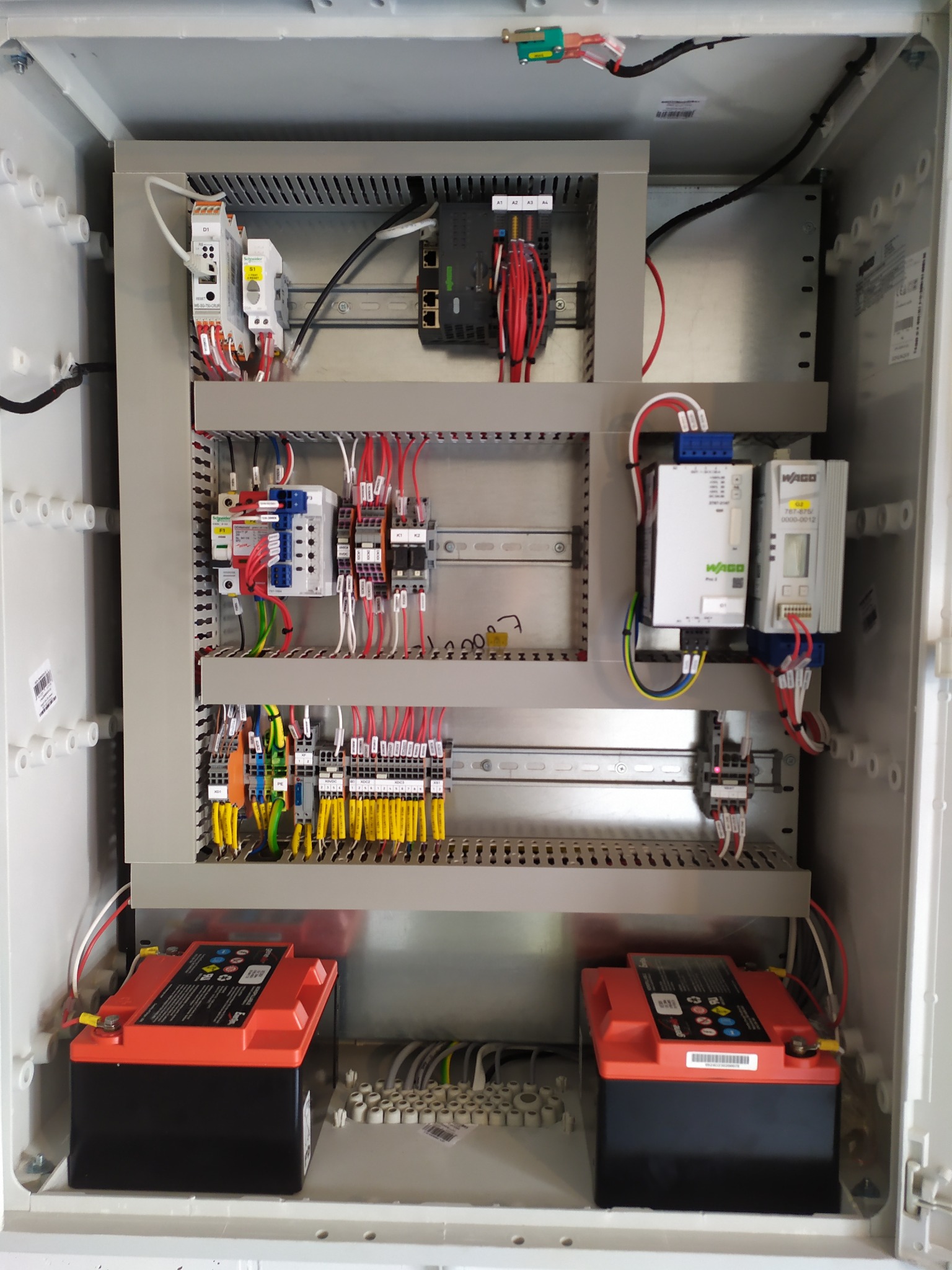 Otwarta szafa sterownicza z akumulatorami, okablowaniem i modułami elektronicznymi. Widoczne złącza, listwy DIN i etykiety. Całość w jasnej obudowie.