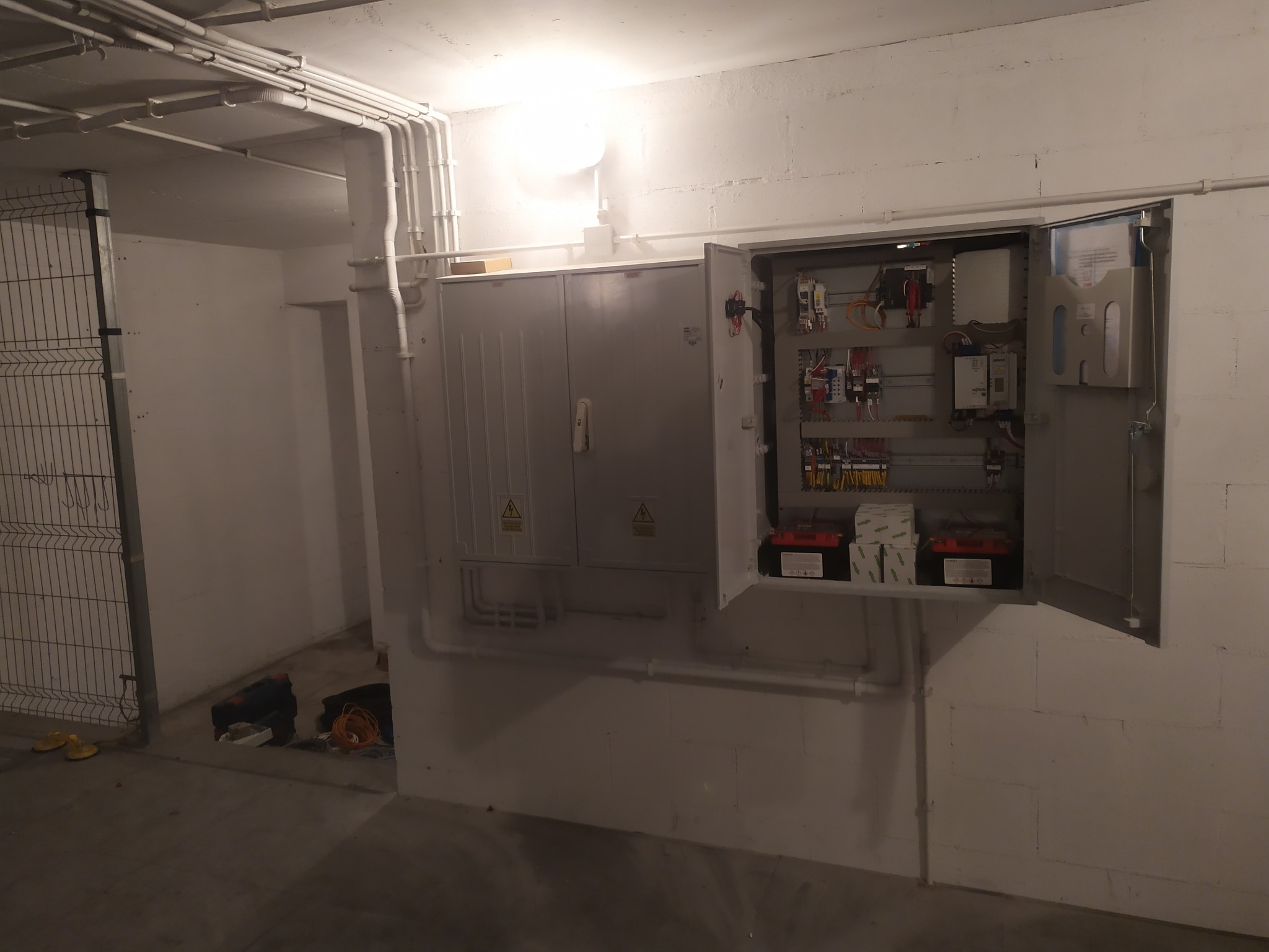 Otwarta szafa sterownicza z widocznymi elementami instalacji elektrycznej, przewody w białych rurkach na ścianie, akumulatory na dolnej półce.
