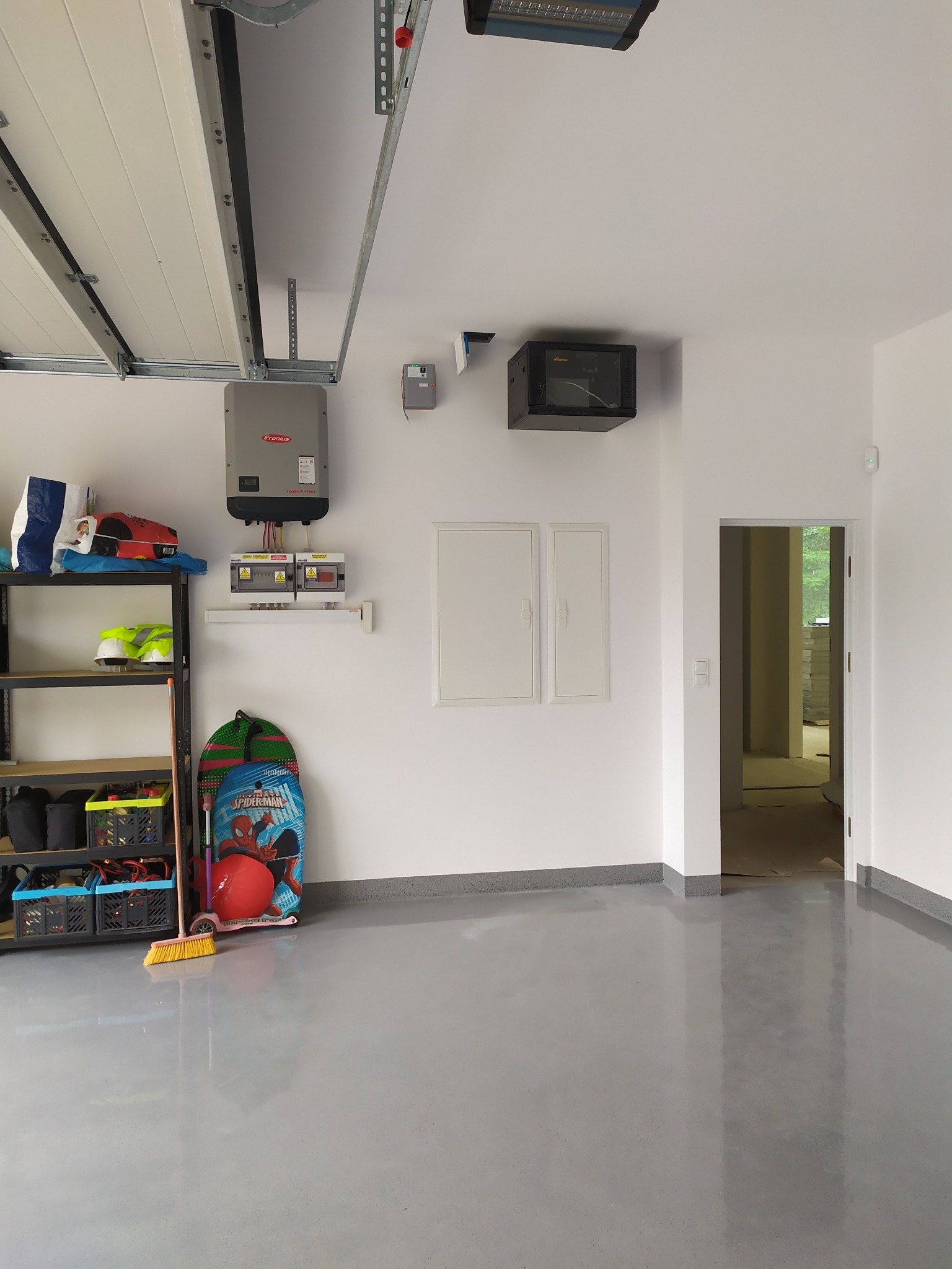 Wykończony garaż z szarą, błyszczącą posadzką żywiczną, regałem, instalacją elektryczną z inwerterem Fronius i szafką RACK na ścianie.