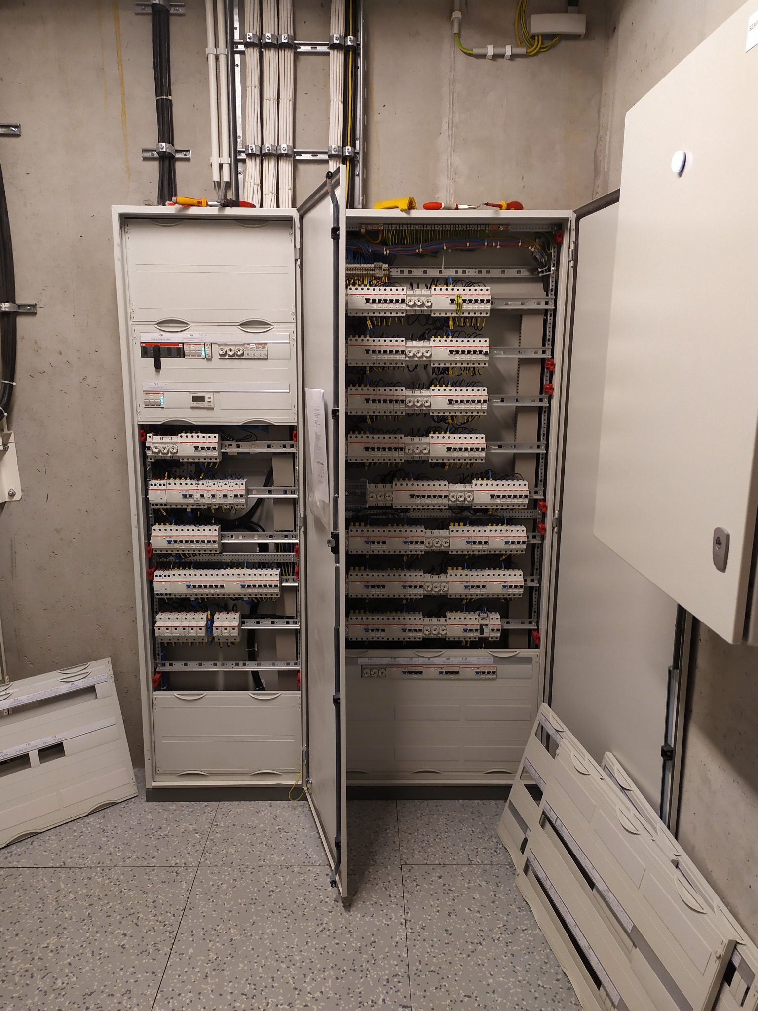 Otwarta szafa elektryczna z widocznymi bezpiecznikami, przewodami i elementami sterującymi. Na górze szafy leżą narzędzia. Widoczne kable w korytach na ścianie.