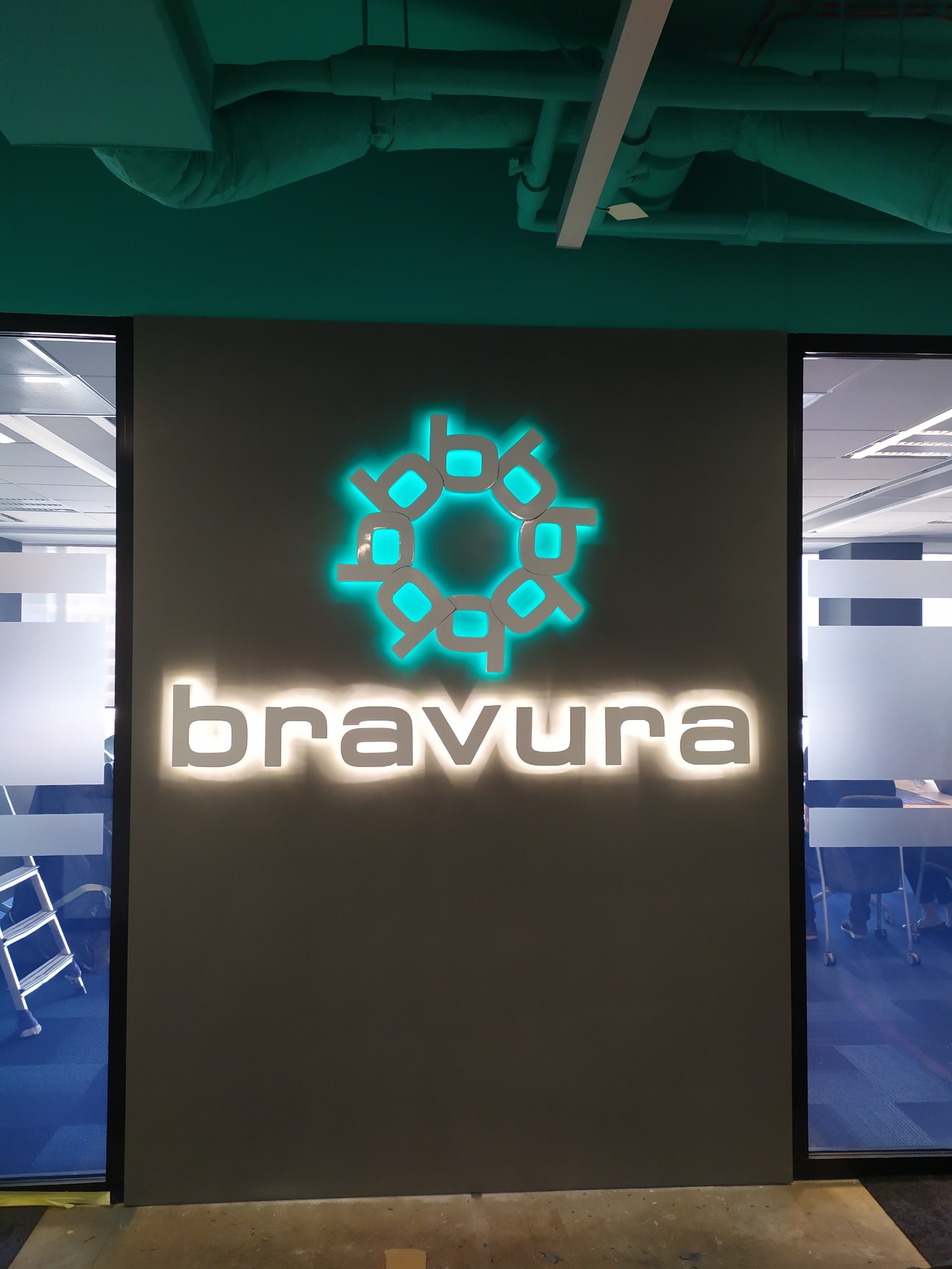 Podświetlane logo 'bravura' na szarej ścianie w biurze. Logo w kolorze turkusowym, nazwa firmy biała. Widoczne rury wentylacyjne pod sufitem i fragmenty biura za szybą.