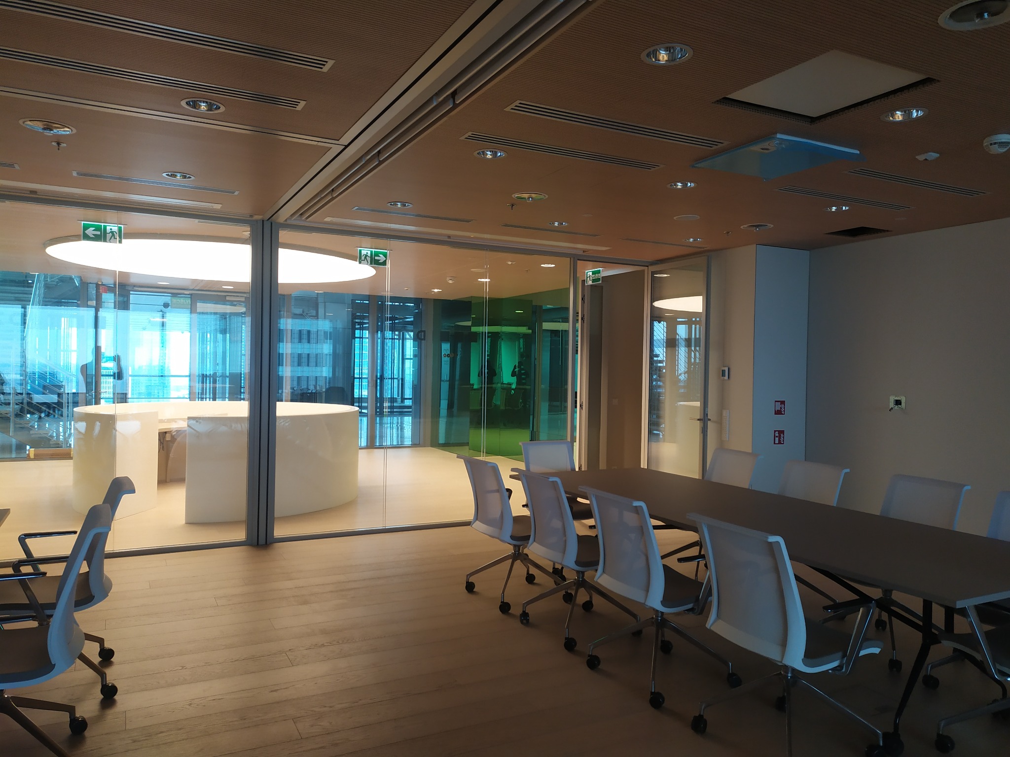 Nowoczesna sala konferencyjna z drewnianą podłogą i sufitem, szklanymi ścianami, oświetleniem sufitowym i stołem z krzesłami. Widoczne znaki ewakuacyjne.