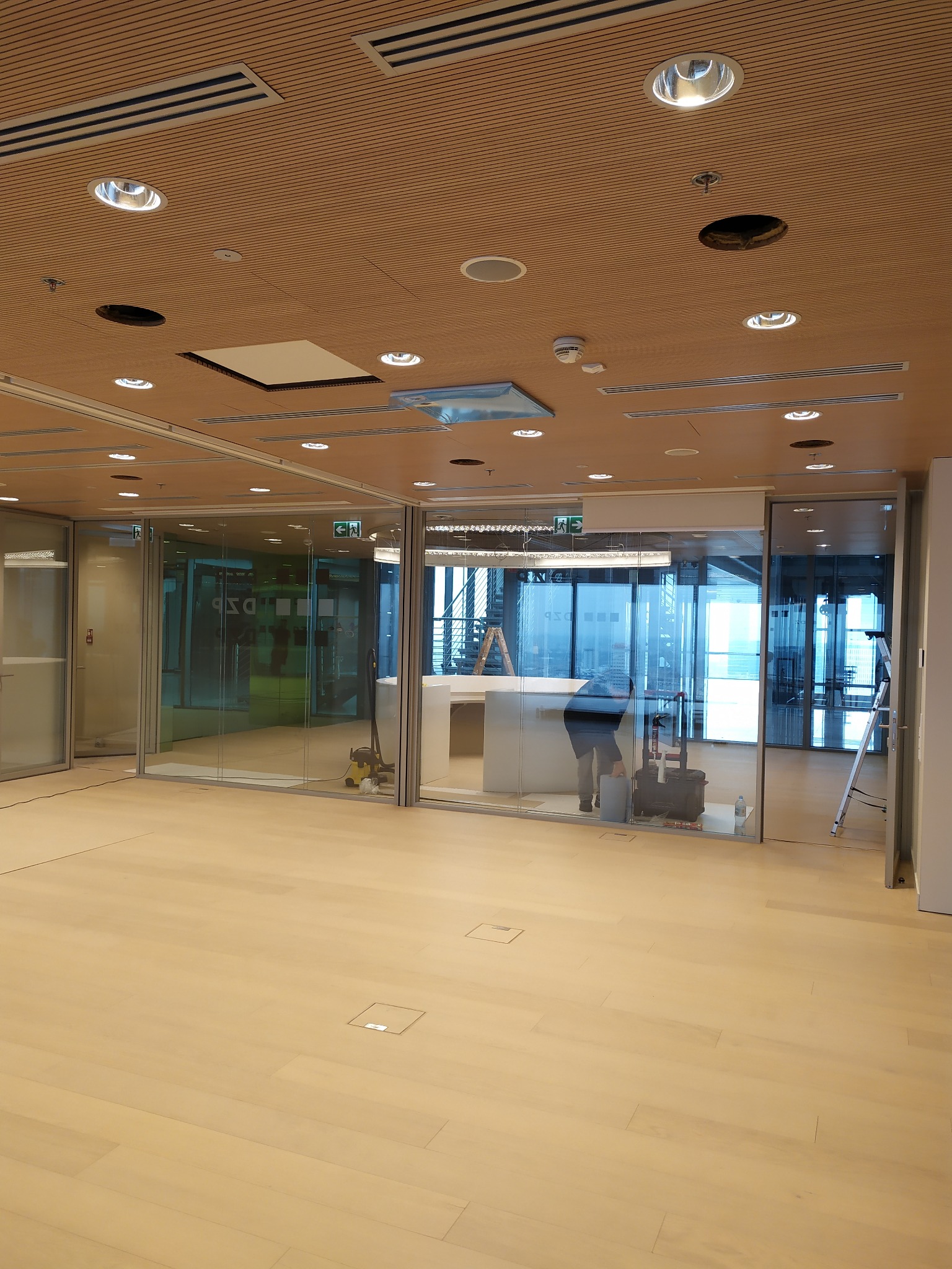 Montaż oświetlenia sufitowego w biurze w Gzy. Widoczne otwory na lampy i pracownik podczas instalacji w przeszklonym pomieszczeniu. Jasna podłoga i drewniany sufit.