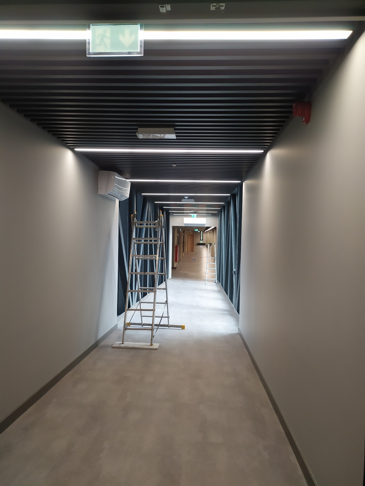 Korytarz biurowy z zamontowanym oświetleniem LED, klimatyzacją i systemem przeciwpożarowym. Widoczna drabina sugeruje trwające prace instalacyjne.