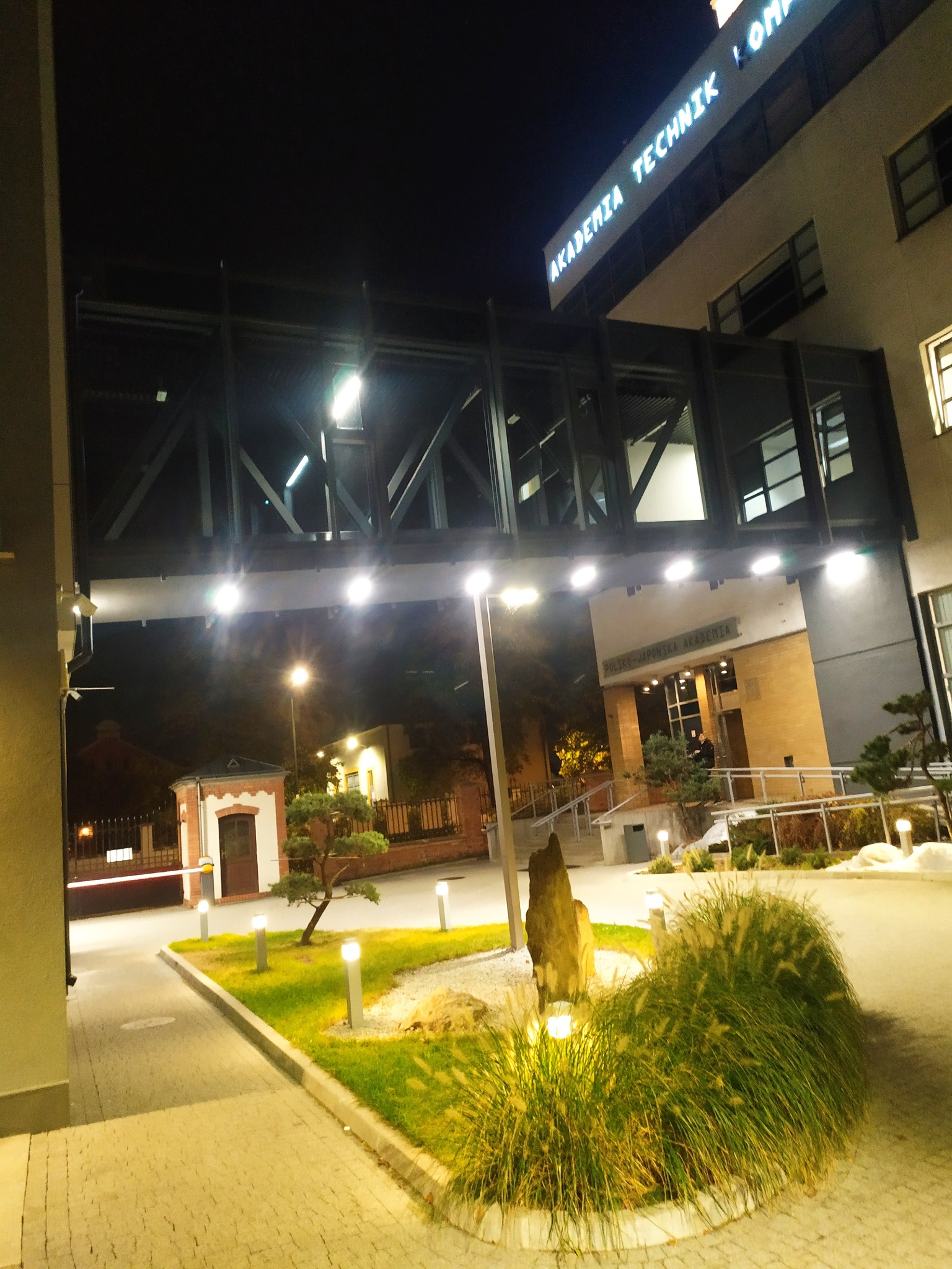 Nocne oświetlenie zewnętrzne budynku uczelni z przeszklonym łącznikiem, lampy parkowe na chodniku i trawniku. Widoczny budynek portierni.