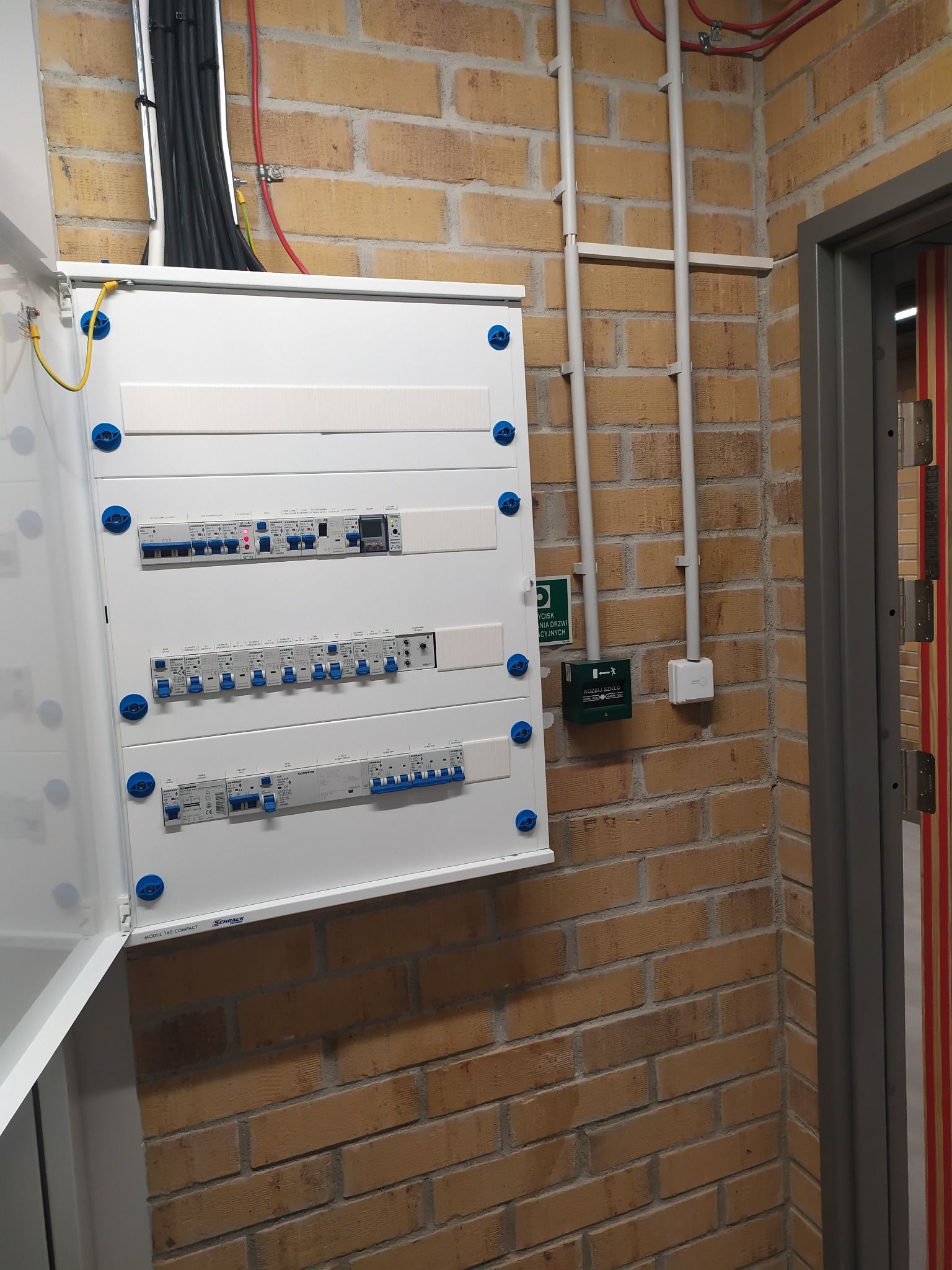 Otwarta, biała skrzynka elektryczna z niebieskimi elementami mocującymi na ceglanej ścianie. Widoczne przewody i zabezpieczenia wewnątrz. Obok drzwi z szarą futryną.
