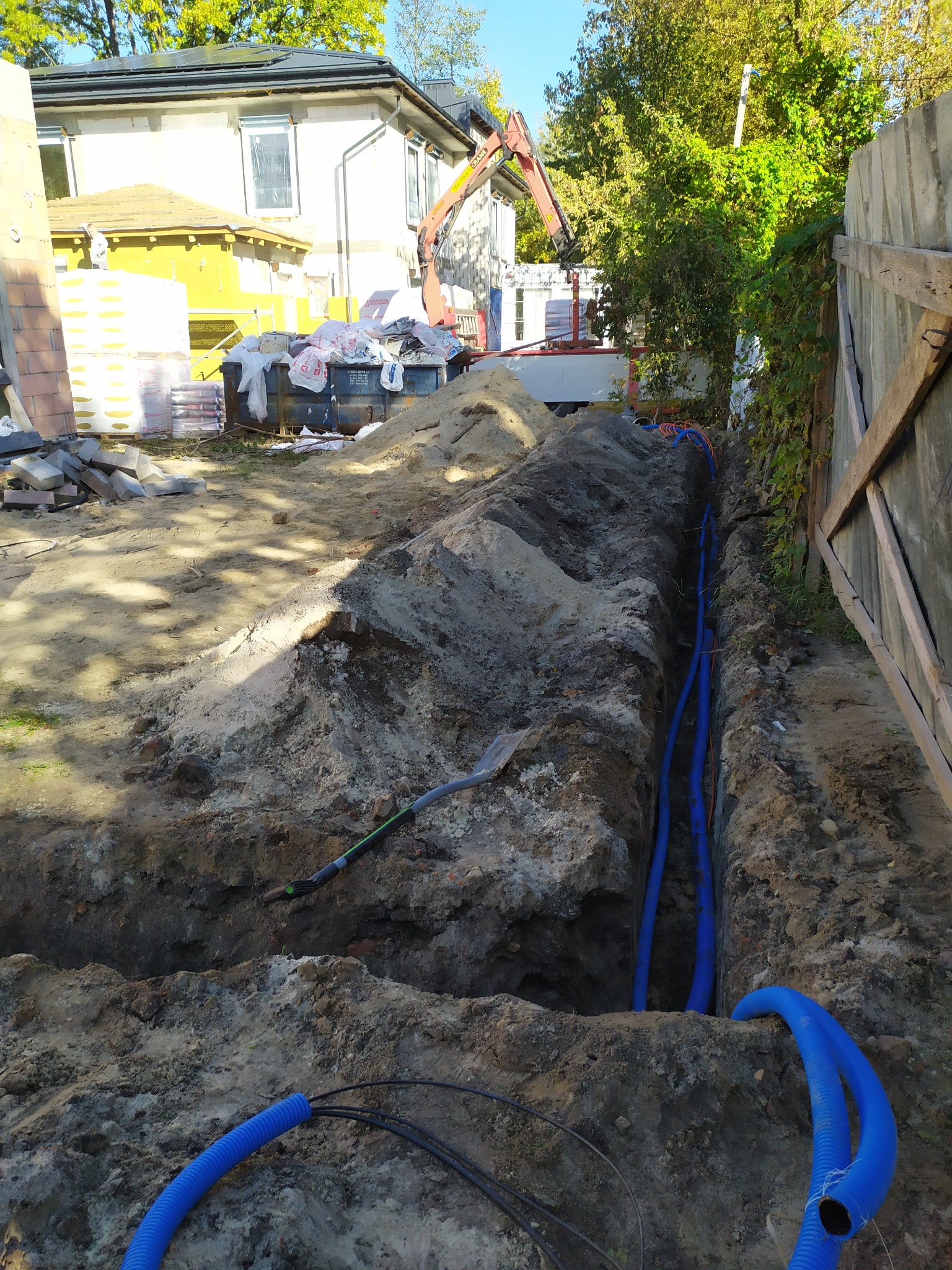 Wykop z niebieskimi rurami kablowymi w ziemi, widok z góry. Na drugim planie budynek w trakcie budowy i sprzęt budowlany. Prace ziemne.