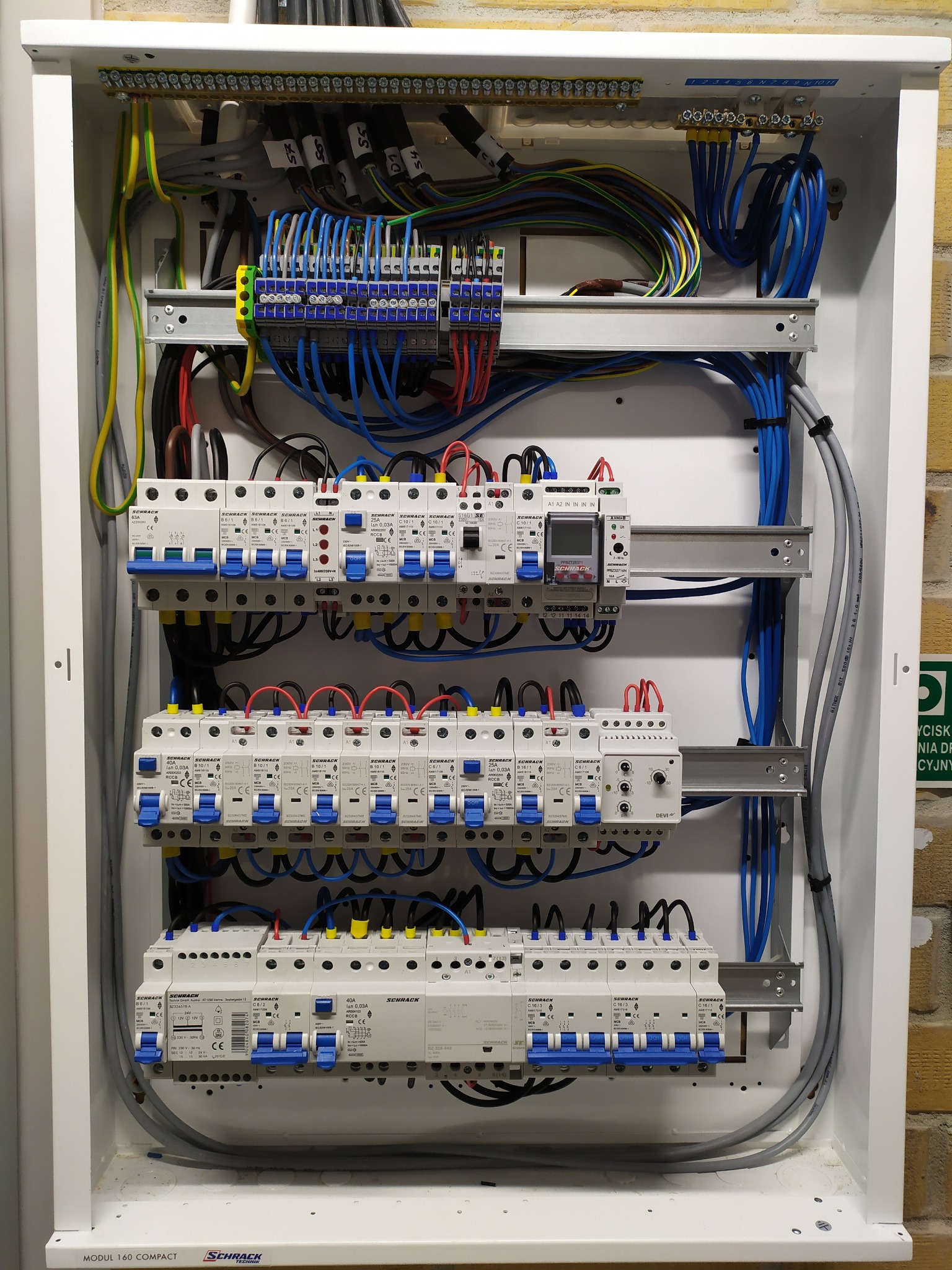 Rozdzielnica elektryczna Schrack z zamontowanymi bezpiecznikami, przewodami i listwami zaciskowymi w białej obudowie, zamontowana na ceglanej ścianie.