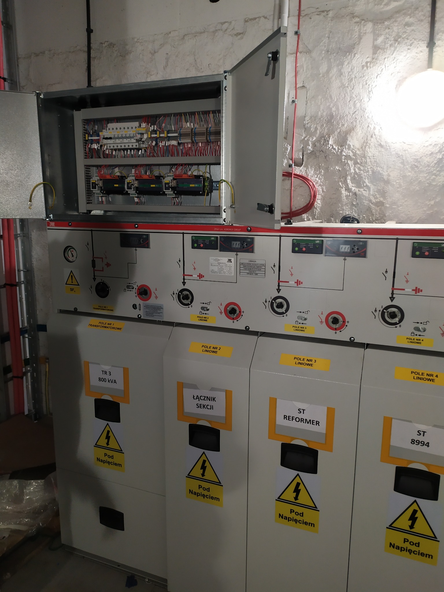 Rozdzielnica elektryczna z otwartą szafą, widoczne kable, bezpieczniki i oznaczenia ostrzegawcze. Białe ściany z surowym wykończeniem w tle.