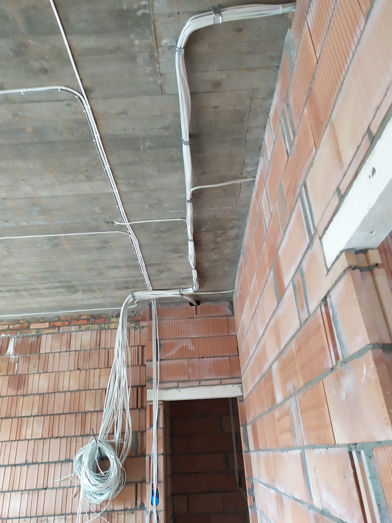 Surowe wnętrze z instalacją elektryczną: przewody w peszlach przymocowane do sufitu i ściany z cegły. W tle widoczne otwory drzwiowe.