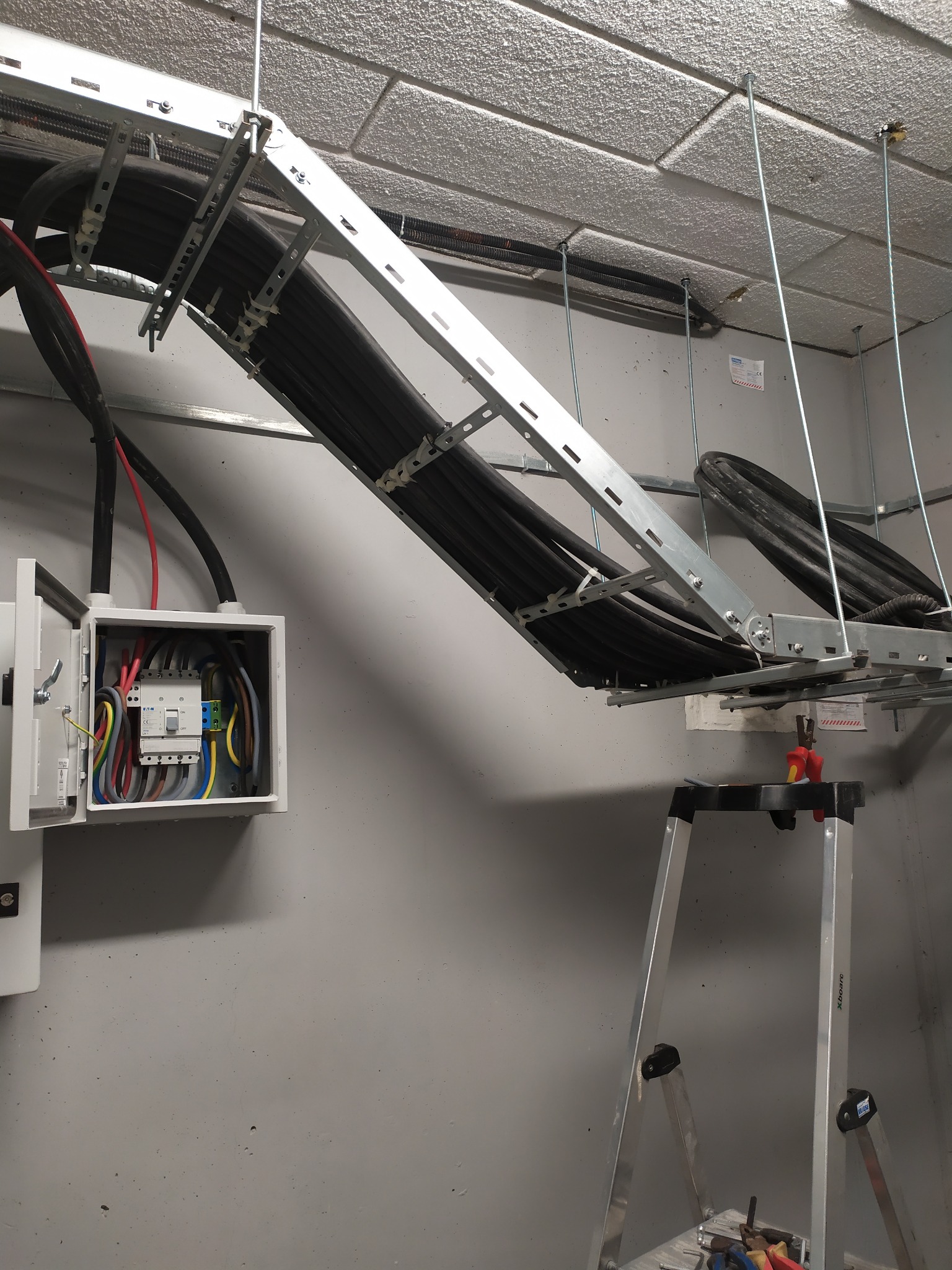 Instalacja elektryczna: kable w korytach zawieszonych pod sufitem, otwarta skrzynka z bezpiecznikami, drabina. Widok na szarą ścianę i sufit z płyt.