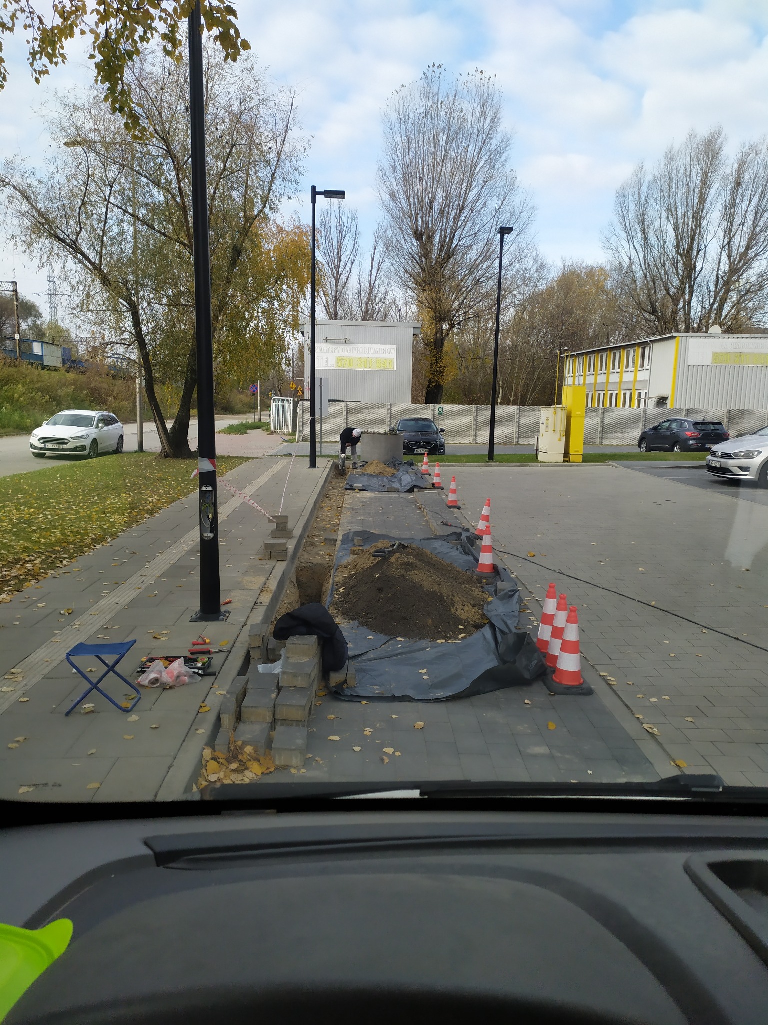 Układanie kostki brukowej przy chodniku, widoczne narzędzia i stożki ostrzegawcze. Prace ziemne w toku, teren zabezpieczony. Jesienne otoczenie, widok z wnętrza pojazdu.