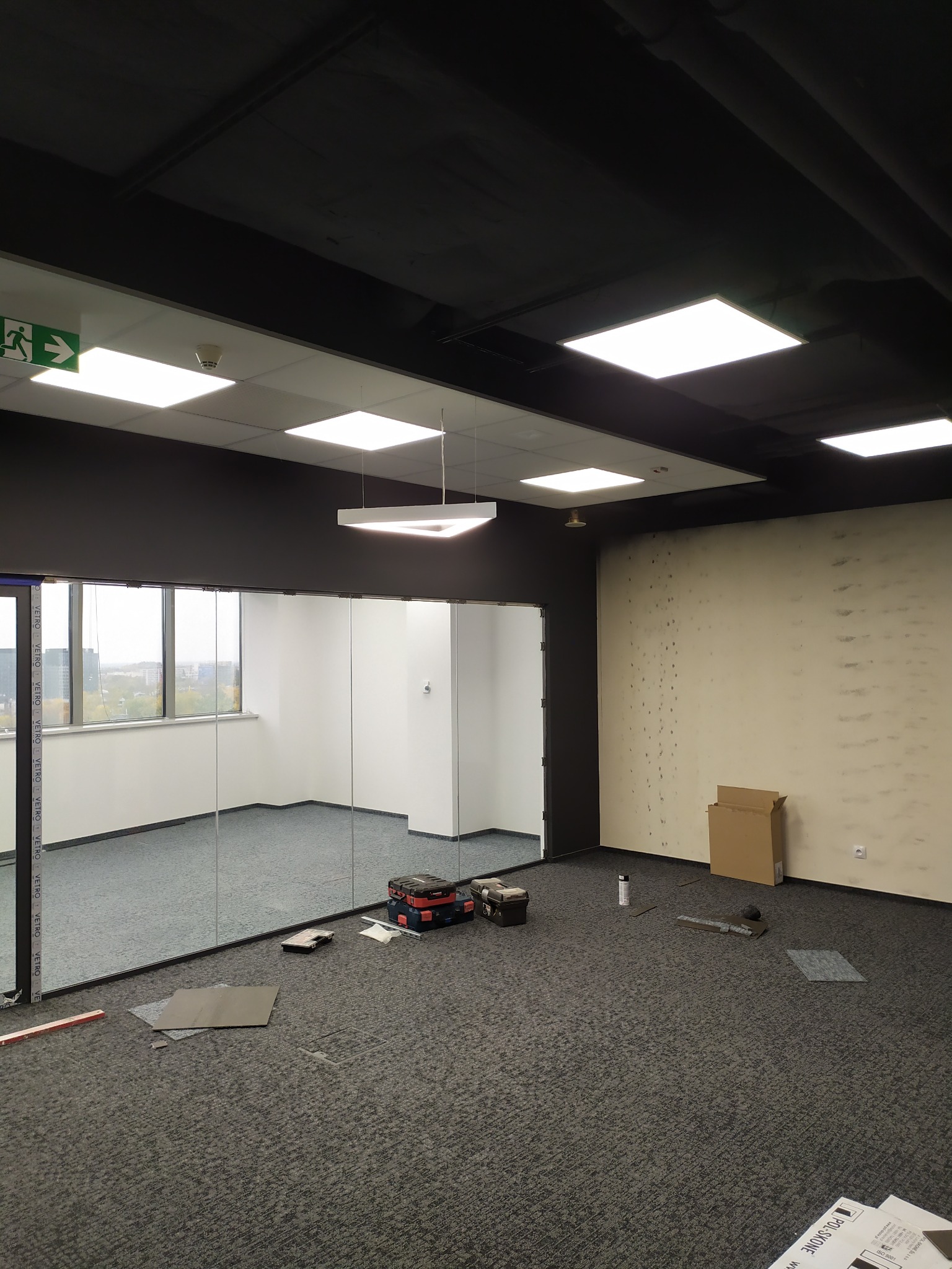 Wnętrze biura w trakcie instalacji oświetlenia: widoczne narzędzia, skrzynka narzędziowa i panele świetlne na suficie. Częściowo zamontowane szklane ścianki działowe.