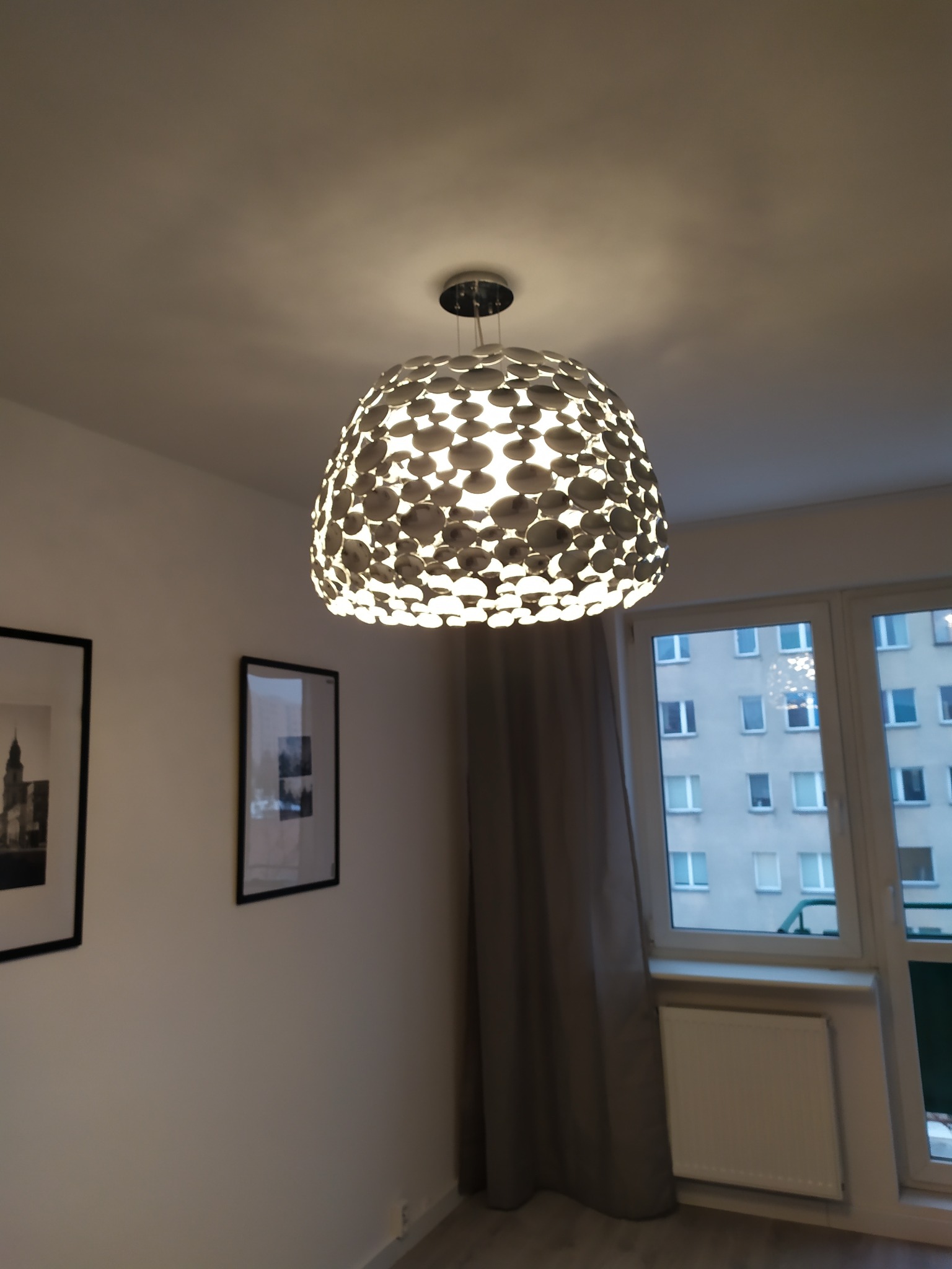 Nowoczesna lampa sufitowa z okrągłymi elementami, oświetlająca wnętrze pokoju z oknem i obrazami na ścianie. Ciepłe światło tworzy przytulną atmosferę.