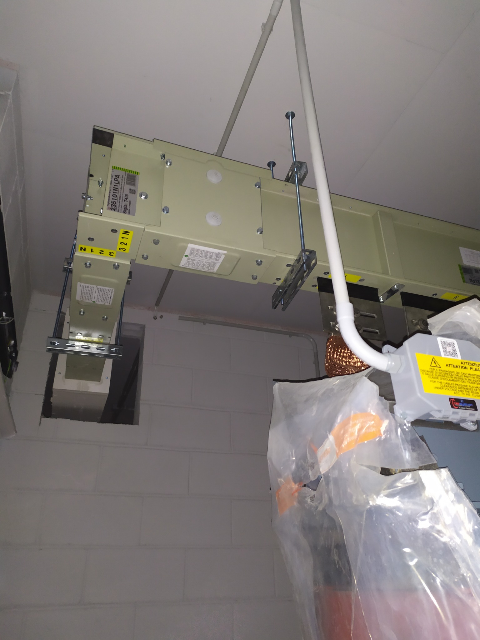 Szyna zbiorcza zasilająca, podwieszona pod sufitem w pomieszczeniu z betonowych bloków. Widoczne elementy mocujące i fragment instalacji elektrycznej.