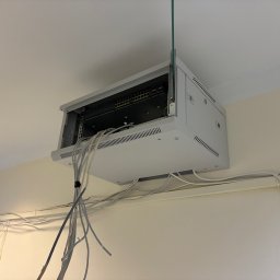 ARTIS STUDIO - ROBERT ZAJĄC - Szafa rack z przełącznikiem sieciowym zamontowana pod sufitem, z widocznymi kablami sieciowymi. Instalacja sieci komputerowej w budynku.