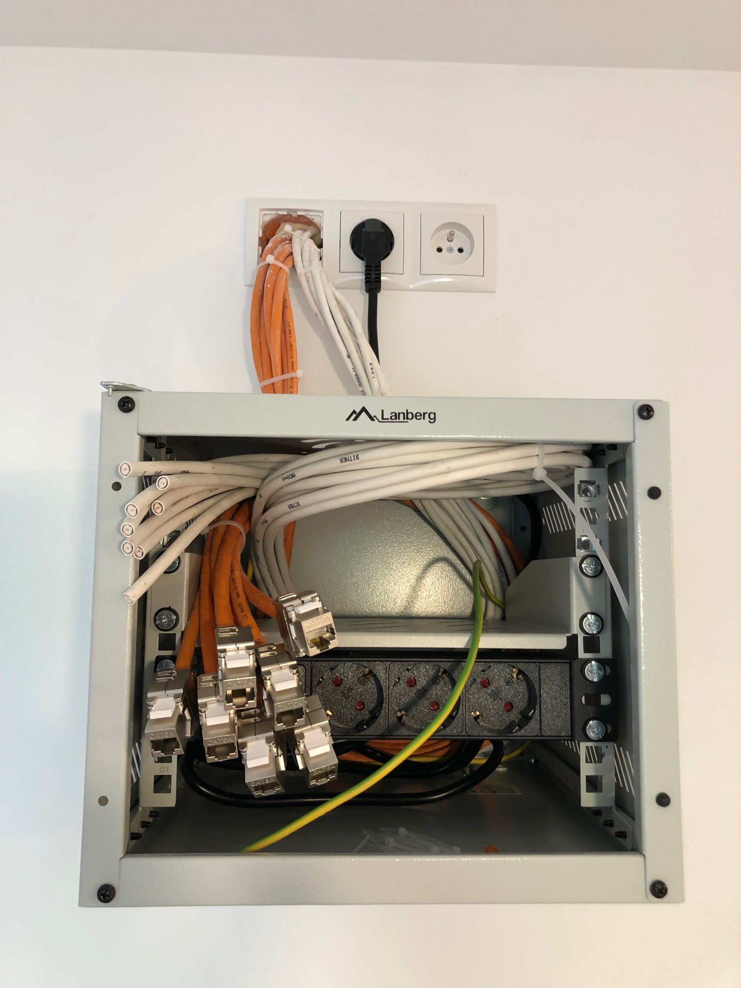 Szary panel krosowniczy Lanberg z kablami sieciowymi (pomarańczowe, białe) i gniazdami zasilającymi zamontowany na białej ścianie.