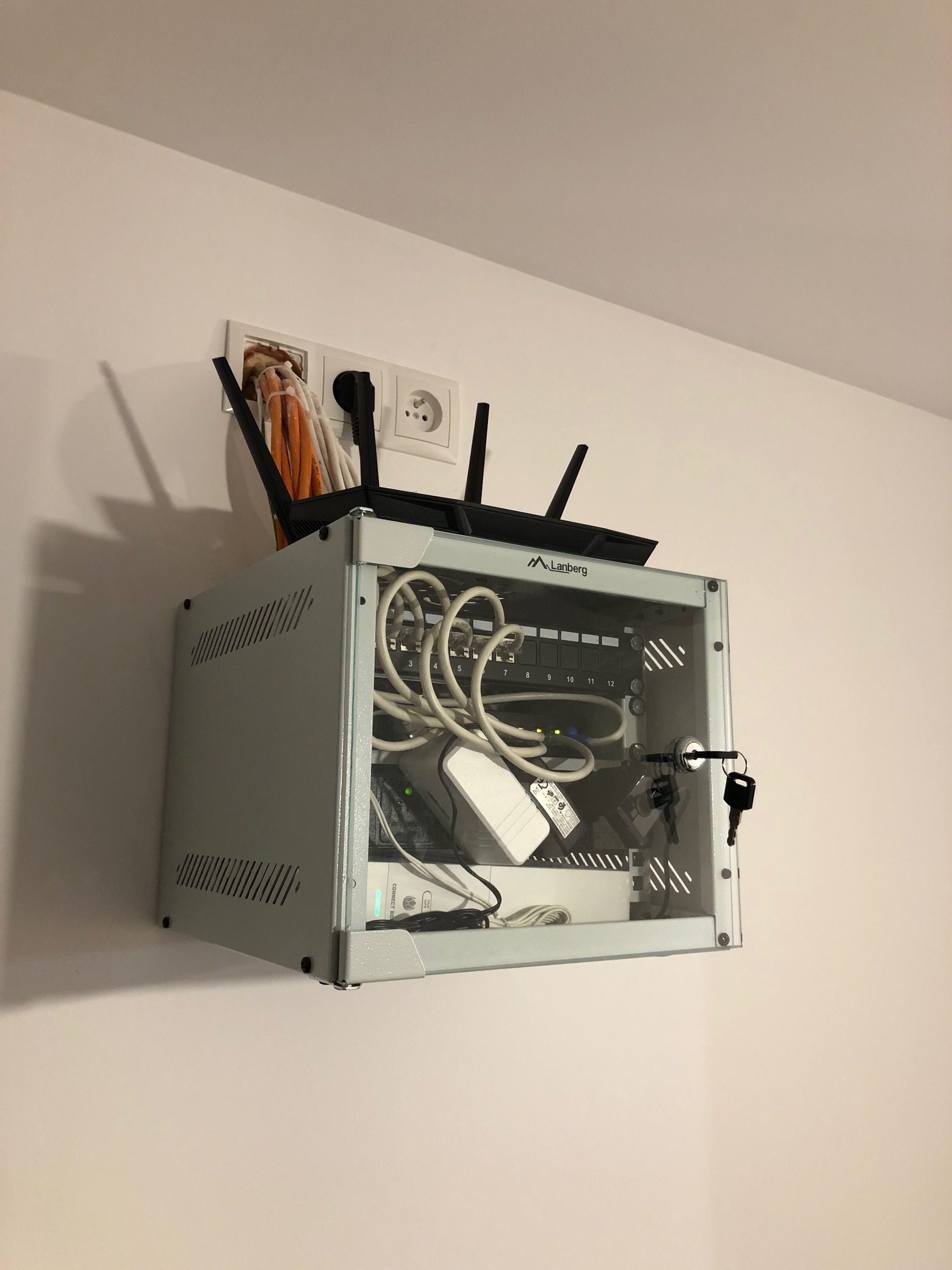 Zamknięta szafka rack Lanberg z routerem, patch panelem i kablami, zamontowana na białej ścianie obok gniazdek elektrycznych. Klucze w zamku.