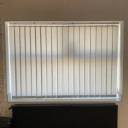 Beżowe, pionowe żaluzje materiałowe w oknie, z widocznym grzejnikiem pod spodem. Stonowana kolorystyka wnętrza.