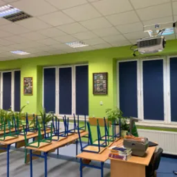 Zasłonięte, granatowe rolety w oknach zielonej sali lekcyjnej. Na suficie widoczny projektor. Krzesła na stołach, doniczki z roślinami na parapetach.