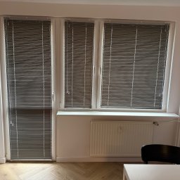 BW Rolety - Szare żaluzje aluminiowe zamontowane w oknach i drzwiach balkonowych. Widoczne białe ramy okienne i jasna ściana z grzejnikiem. Parkiet ułożony w jodełkę.