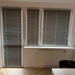 Szare żaluzje aluminiowe zamontowane w oknach i drzwiach balkonowych. Widoczne białe ramy okienne i jasna ściana z grzejnikiem. Parkiet ułożony w jodełkę.
