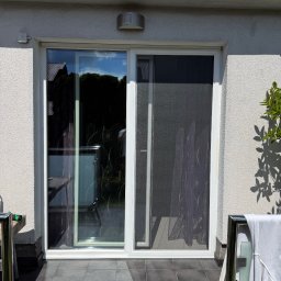 BW Rolety - Przesuwne drzwi balkonowe z moskitierą w białej ramie, widok z zewnątrz na taras z szarymi płytkami. Cień drzewa na ścianie budynku.