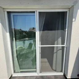 Białe, przesuwne drzwi balkonowe z moskitierą na tle jasnej elewacji. Widoczny fragment balkonu i odbicie otoczenia w szybie.