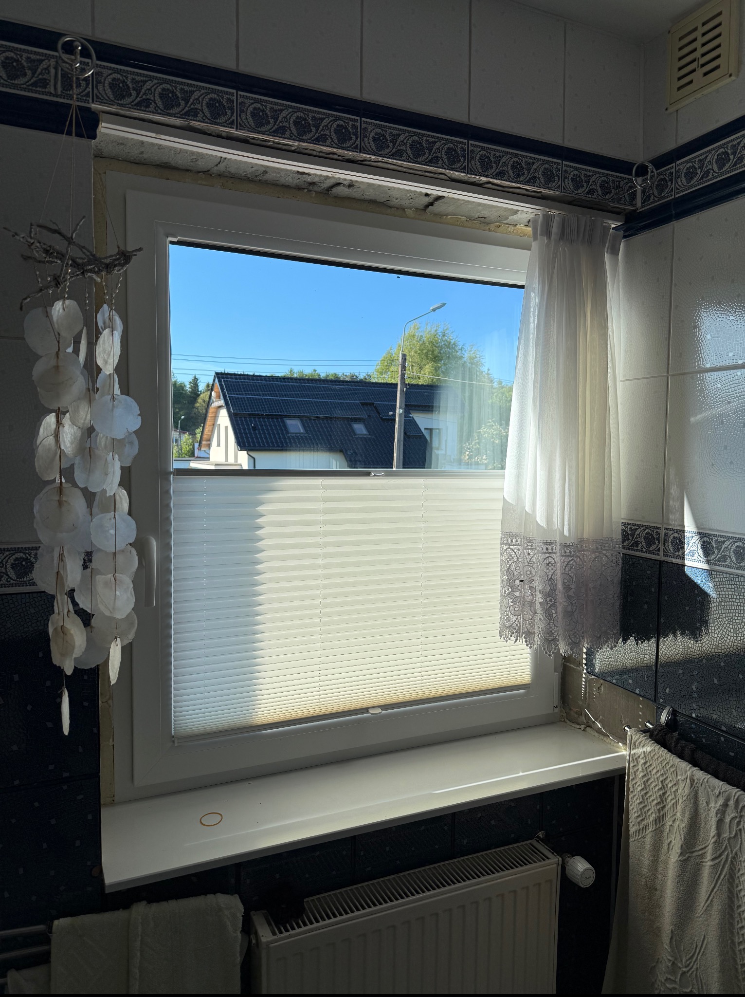 Biała plisa okienna w łazience z dekoracyjnymi płytkami i widokiem na dom z czarnym dachem. Słońce oświetla wnętrze, tworząc jasne refleksy na powierzchniach.