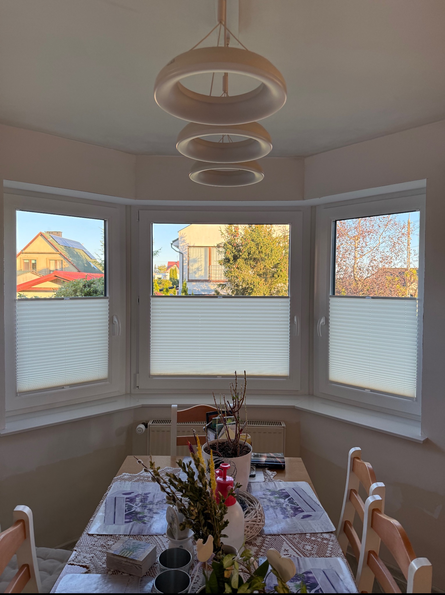 Trzy okna wykuszowe z białymi plisami, widok na domy i drzewa. Na stole dekoracje wiosenne, nad oknami lampa pierścieniowa. Jasne, naturalne światło.