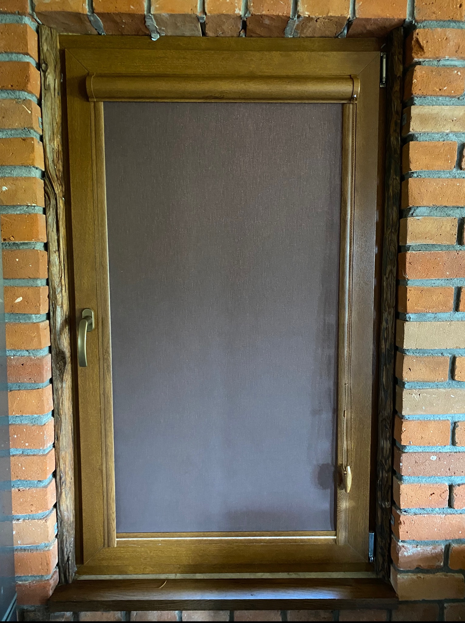 Brązowa roleta wewnętrzna w drewnianej ramie okiennej, zamontowana w murze z cegły. Widoczny mechanizm łańcuszkowy i uchwyt okienny.