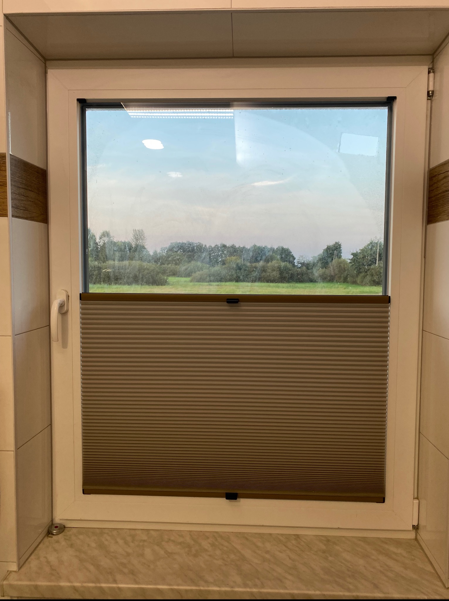 Beżowa plisa okienna zamontowana w białym oknie PCV. Zza szyby widok na zielony krajobraz. Plisa częściowo zasłonięta, widoczne poziome fałdy materiału.