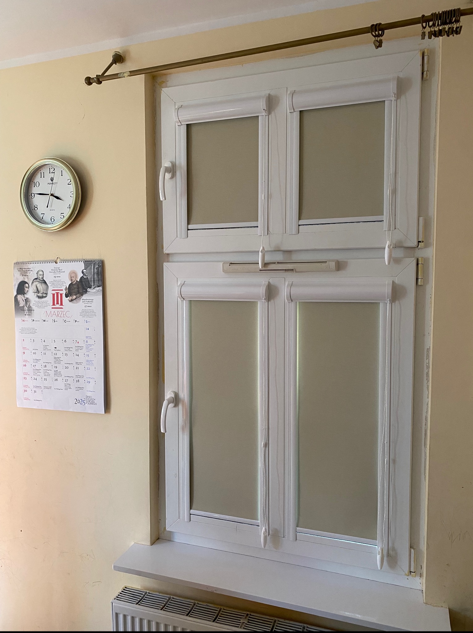 Białe okno z zamontowanymi roletami wewnętrznymi w kolorze beżowym, widok z przodu, z kalendarzem i zegarem na ścianie. Detal wykończenia wnętrza.