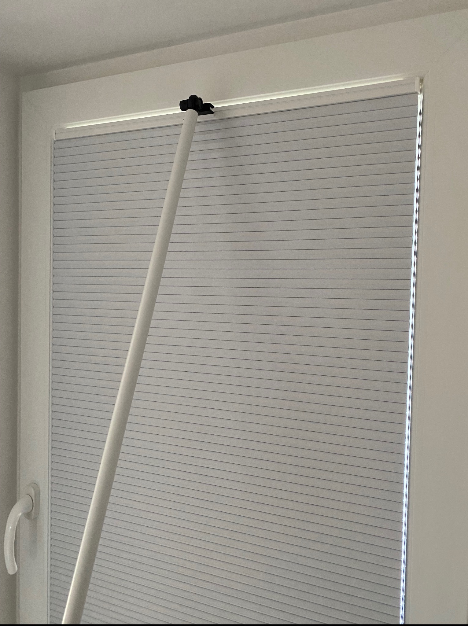 Zamknięte, jasnoszare plisy okienne w białej ramie okiennej, z widocznym białym drążkiem do sterowania na tle białej ściany.