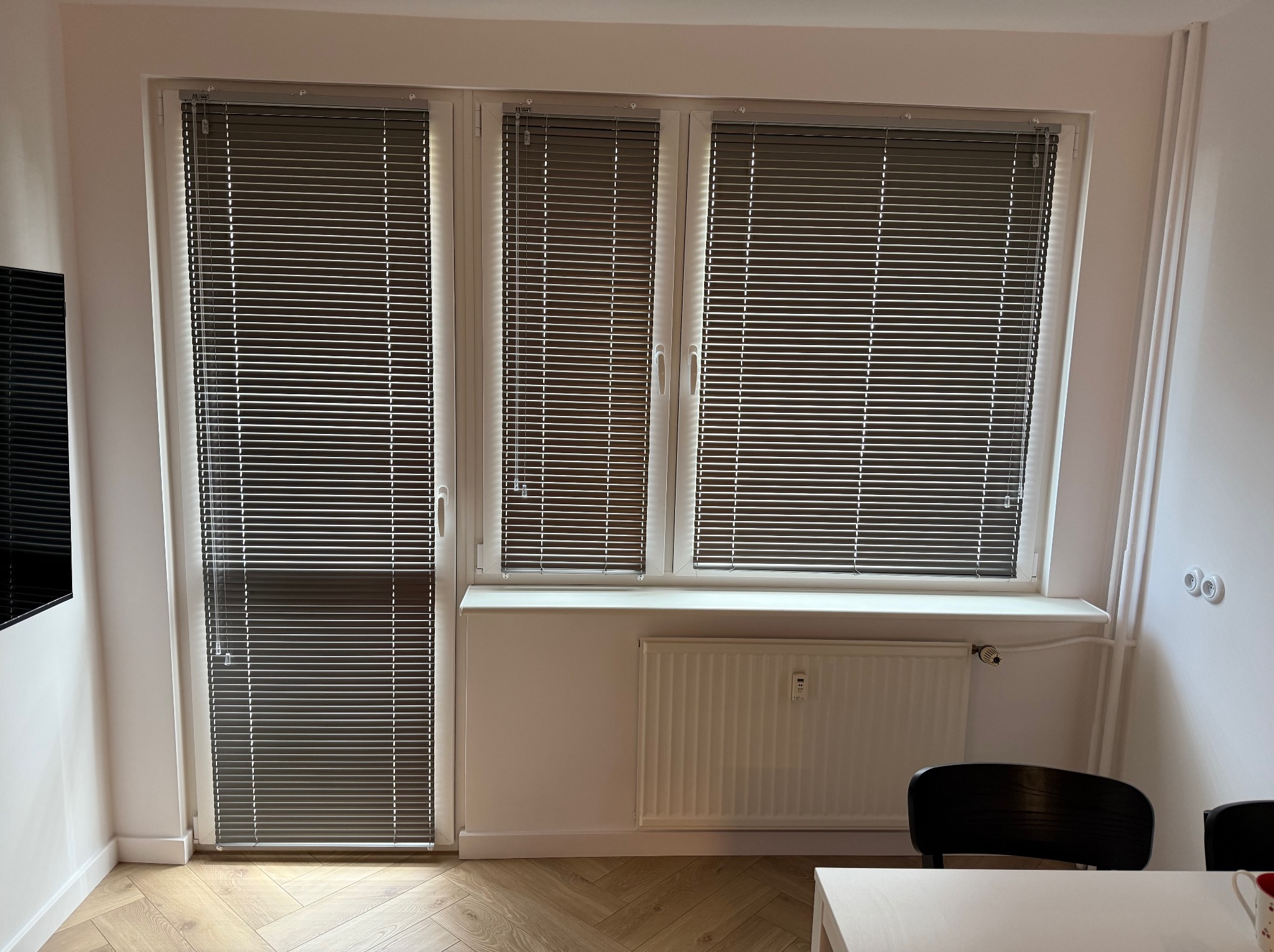 Szare żaluzje aluminiowe zamontowane w oknach i drzwiach balkonowych. Widoczne białe ramy okienne i jasna ściana z grzejnikiem. Parkiet ułożony w jodełkę.