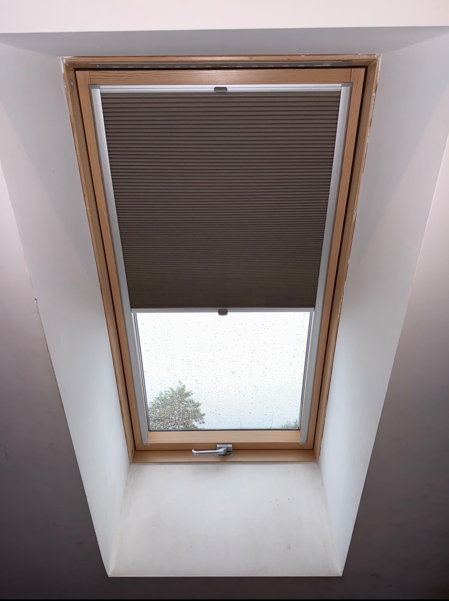Beżowa plisa zamontowana w oknie dachowym z drewnianą ramą. Za oknem widoczny deszczowy krajobraz z drzewem.