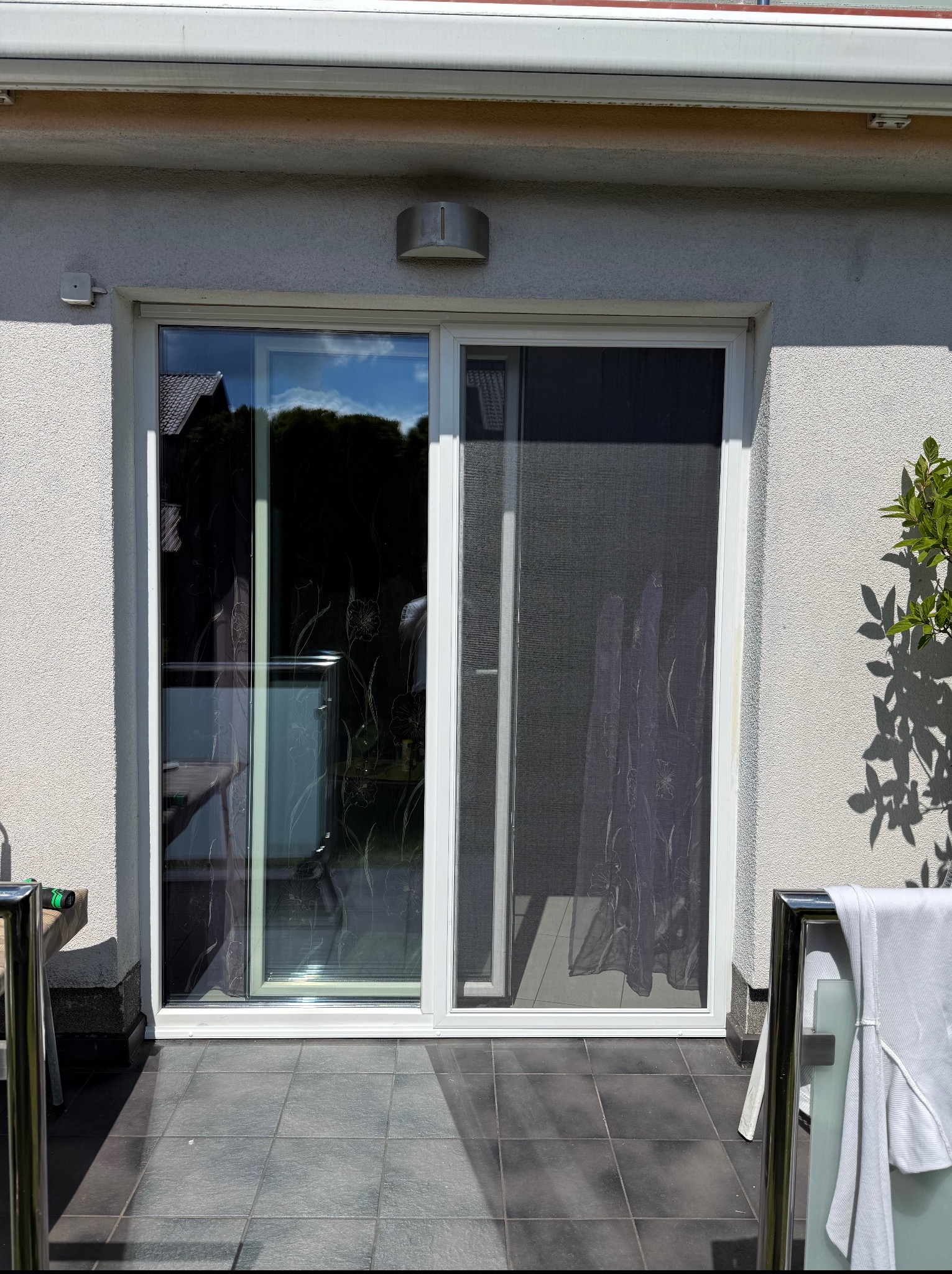 Przesuwne drzwi balkonowe z moskitierą w białej ramie, widok z zewnątrz na taras z szarymi płytkami. Cień drzewa na ścianie budynku.
