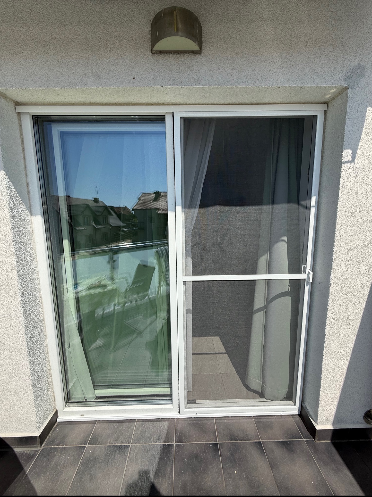 Białe, przesuwne drzwi balkonowe z moskitierą na tle jasnej elewacji. Widoczny fragment balkonu i odbicie otoczenia w szybie.