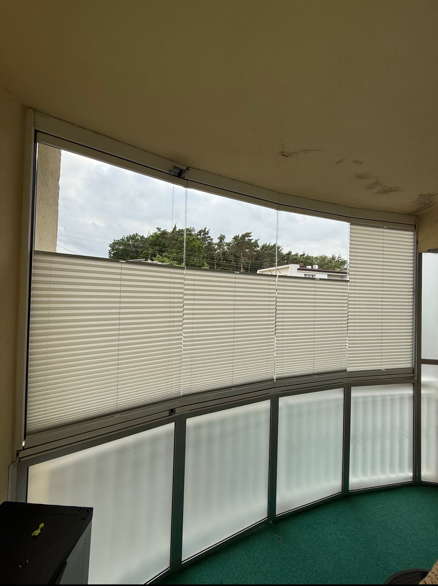 Jasne plisy okienne zamontowane na balkonie z widokiem na zieleń i budynek. Uwagę zwraca nietypowy, zaokrąglony kształt zabudowy balkonu.