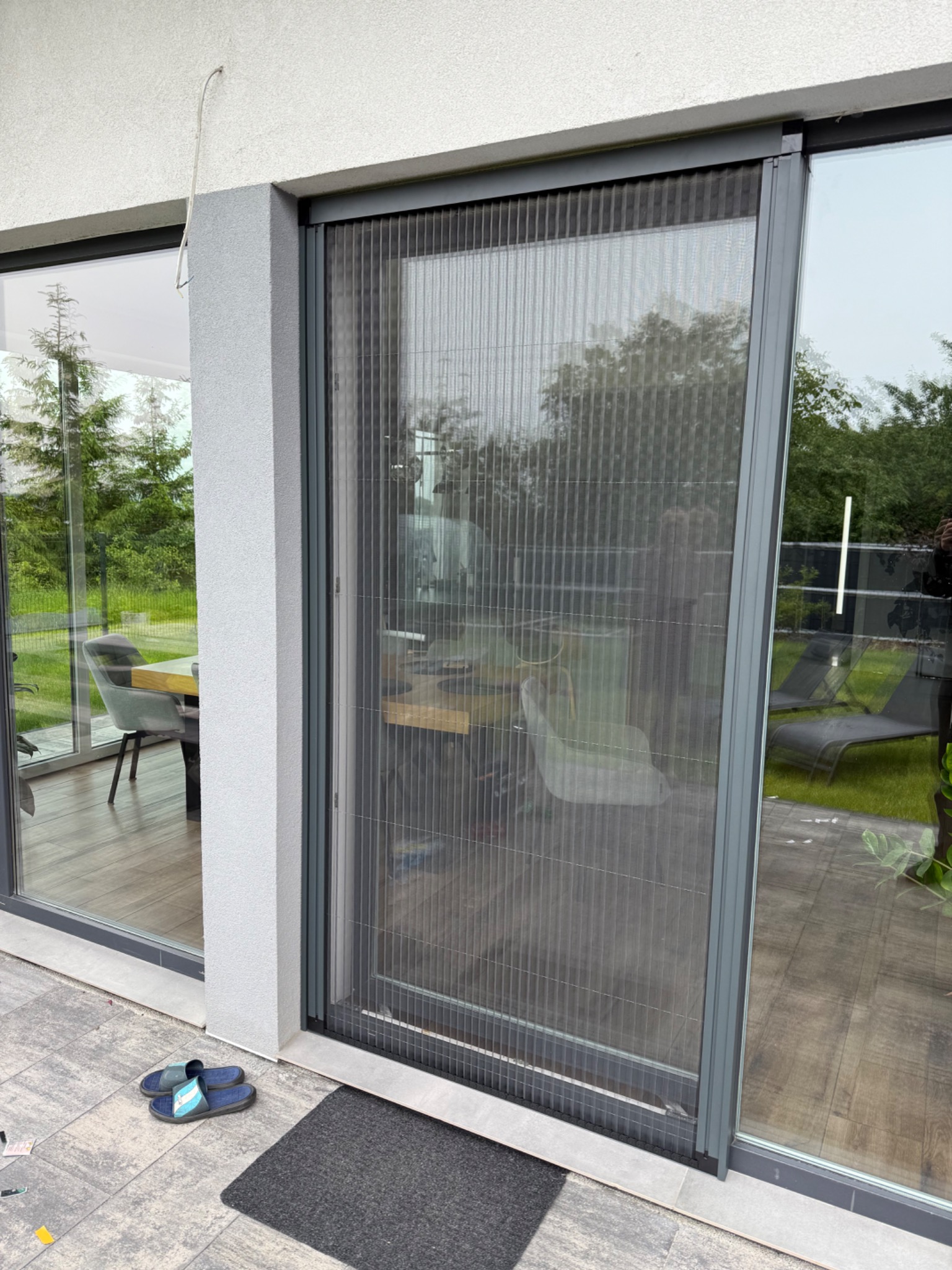 Zewnętrzna moskitiera plisowana w kolorze szarym, zamontowana na tarasowych drzwiach przesuwnych. Widoczne detale konstrukcyjne i otoczenie domu.