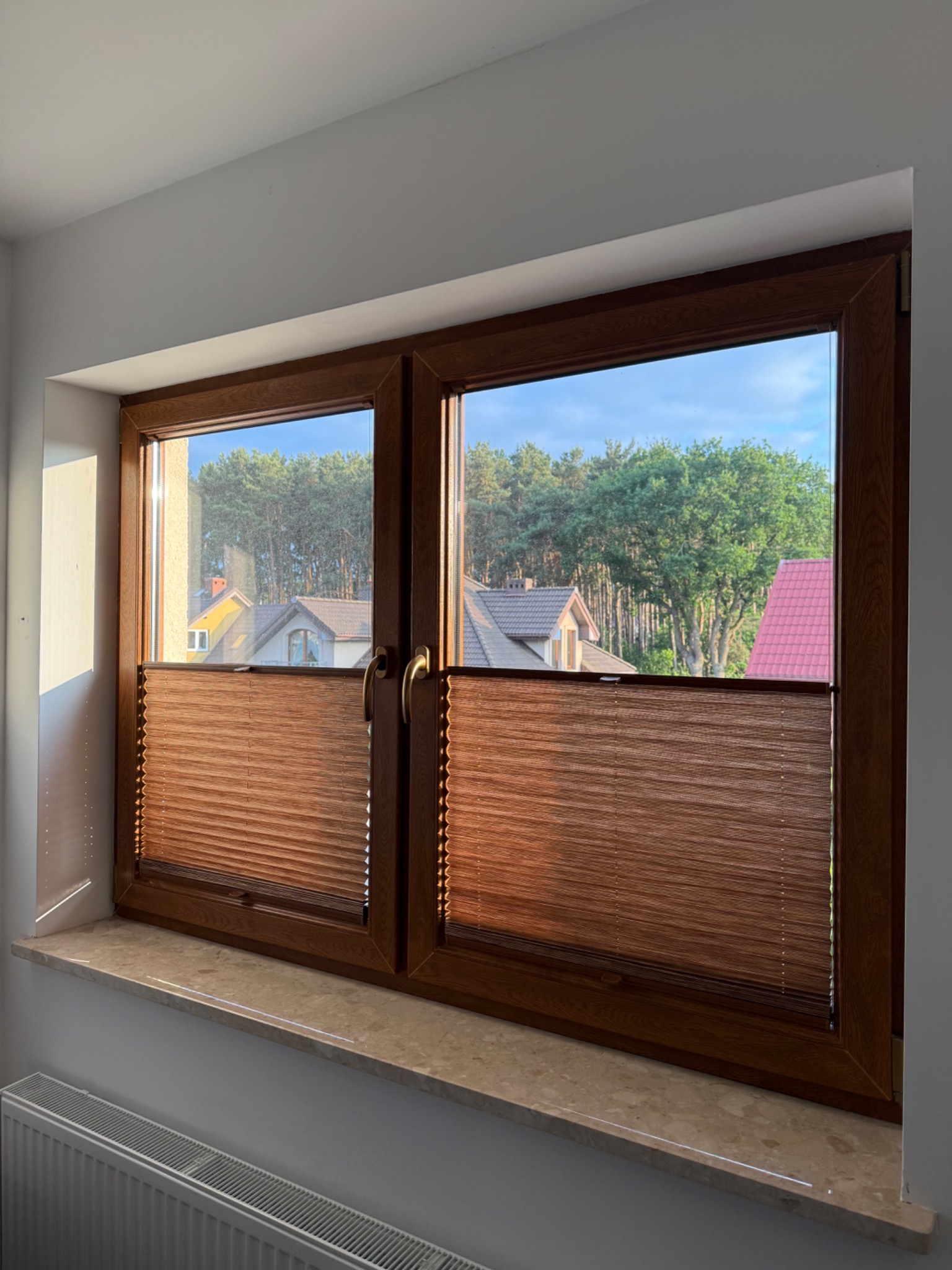 Drewniane okno z brązowymi plisami, częściowo zasłoniętymi, z widokiem na domy i drzewa. Promień słoneczny na ścianie.