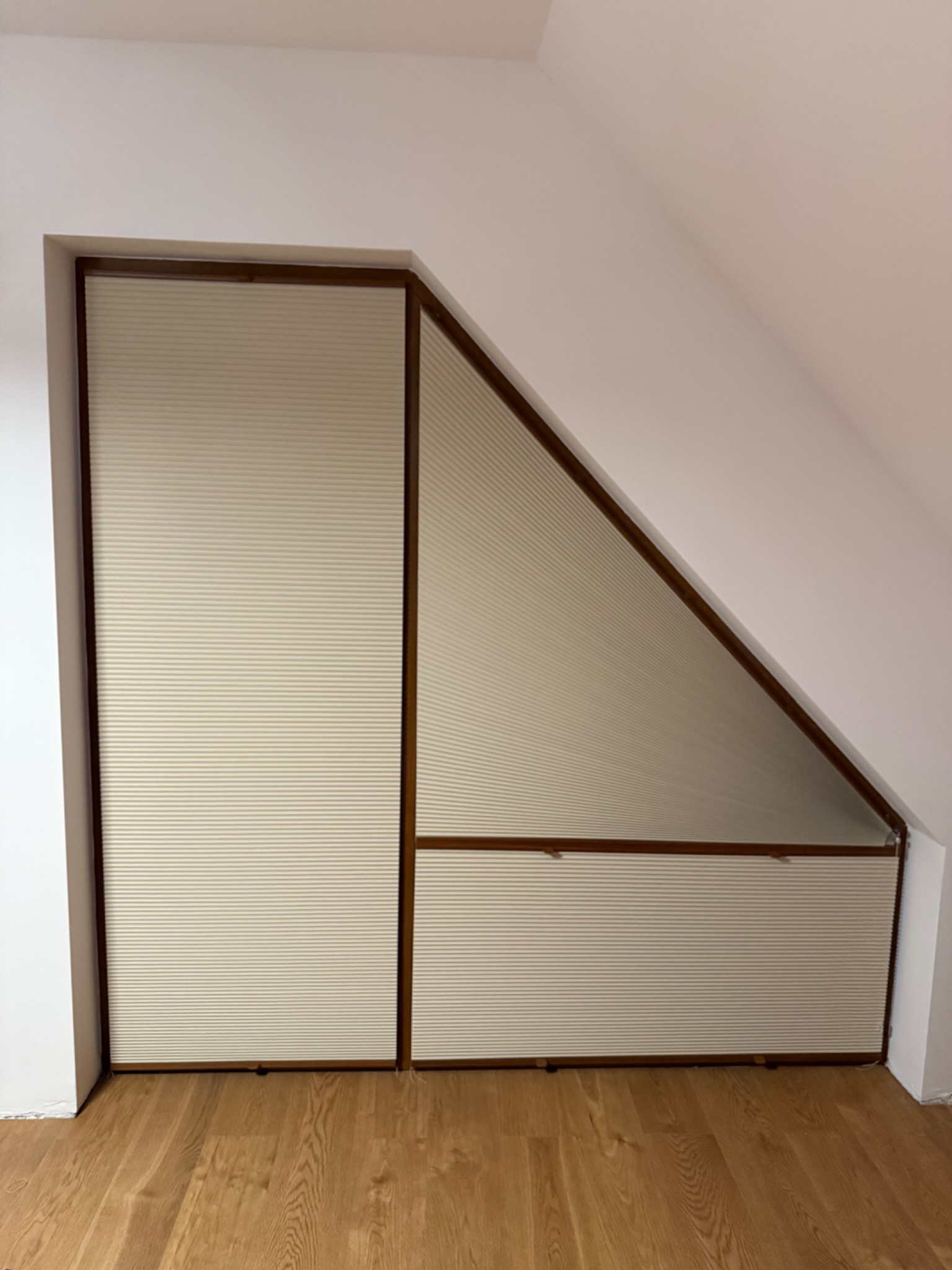 Beżowe plisy okienne w drewnianej ramie, dopasowane do skosu poddasza. Widok na drewnianą podłogę.