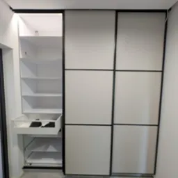 Biała szafa wnękowa z przesuwnymi drzwiami i czarnymi ramami. Jedna część szafy otwarta, widoczne półki i wysuwana szuflada z ubraniami. Szara podłoga.